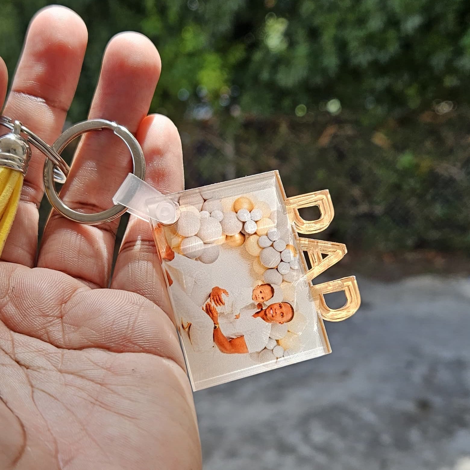 Hand holding a keychain with 'DAD' and a balloon design against a blurred natural background